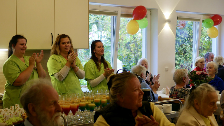 Drei stehende Pflegekräfte in grüner Arbeitskleidung und sitzende Gäste klatschen im bunt geschmückten Saal bei der Wiedereröffnung der Tagespflegestätte für ältere Menschen im Immanuel Seniorenzentrum Kläre Weist in Petershagen
